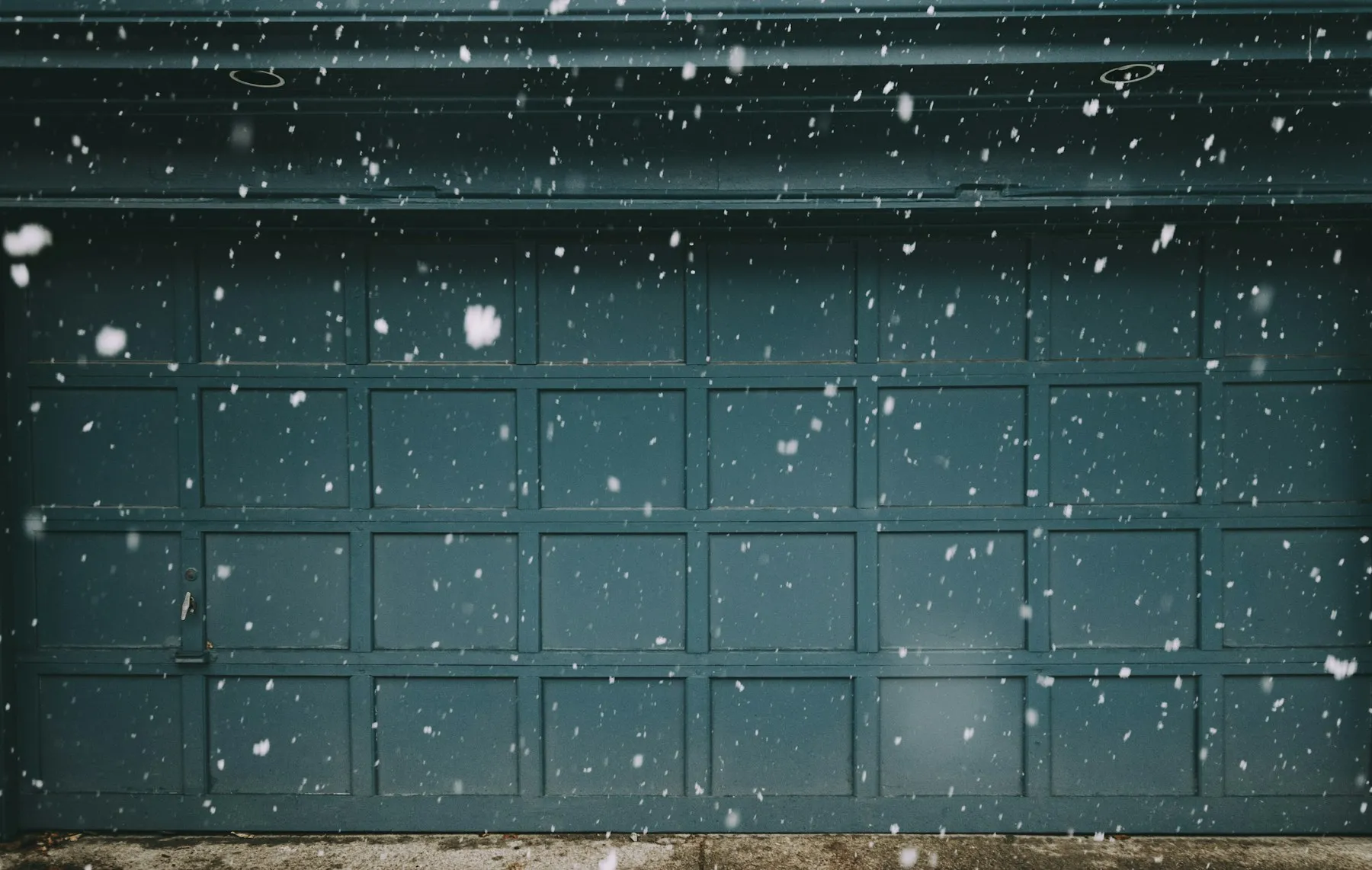A teal raised-panel garage door with snow falling